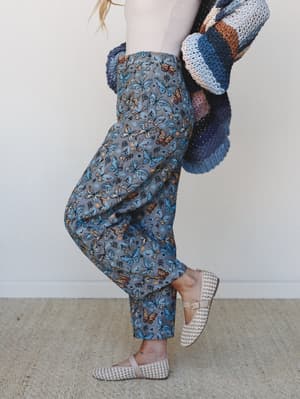 A side-angle lifestyle shot featuring wide-leg butterfly print pants in moody grey paired with woven flats and a chunky color-block crochet cardigan draped over the arm — the kind of outfit that looks like it came together effortlessly but definitely