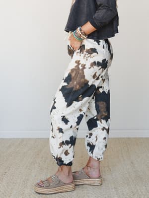 A side-profile shot of cow print jogger pants styled with a cropped charcoal top, stacked beaded bracelets, and espadrille platform sandals — relaxed and a little wild in the best way.
