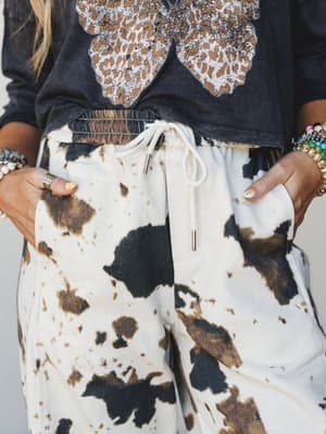 A close-up torso shot of cow print drawstring pants paired with a washed charcoal graphic tee, styled with stacked beaded bracelets and rings for a relaxed western-boho feel.