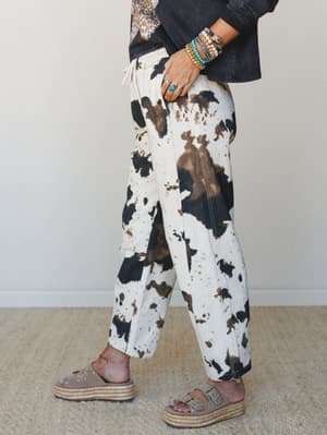 A side-profile shot of cow print relaxed pants styled with a washed black jacket, layered beaded bracelets, and platform espadrille sandals — the kind of outfit that looks effortless but definitely has a point of view.