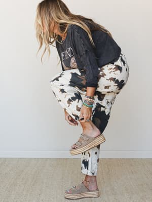 A model bends down to adjust her platform sandal, showing off cow print pants paired with a washed graphic tee and a stack of beaded bracelets — the kind of effortlessly collected outfit that looks like it came together in five minutes and took zero
