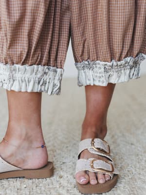 A close-up detail shot of wide-leg gingham pants with a contrast ruffle hem, paired with double-buckle slide sandals and a delicate evil eye anklet — the kind of effortless summer outfit that looks like it happened on purpose.