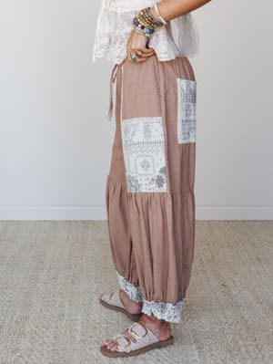 A side-profile shot of relaxed harem-style pants in a warm caramel gingham check, styled with a white lace top, stacked beaded bracelets, and buckle sandals — the kind of outfit that looks like it was effortlessly assembled but definitely wasn't.