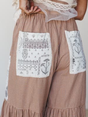 A close-up detail shot of wide-leg gingham pants in a warm caramel tone, featuring oversized patch pockets with intricate botanical and folk print detailing and crochet trim edging — paired with a sheer lace crop top peeking into frame.