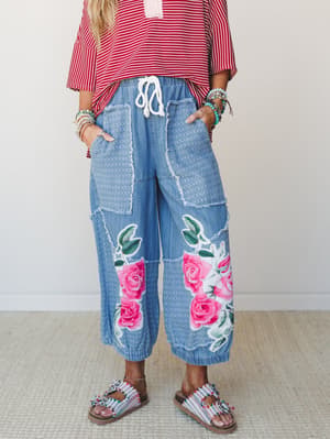 Wide-leg denim pants with bold rose appliqués and patchwork detailing styled with a red striped top, beaded sandals, and stacked turquoise bracelets — the kind of outfit that makes a statement without trying too hard.