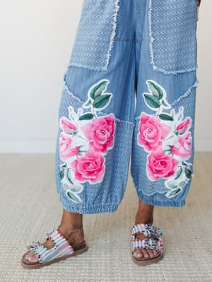 A close-up of wide-leg denim pants in light blue with mixed textures and patchwork panels, anchored by bold pink rose appliqués on each leg — styled with beaded embellished sandals on a natural woven rug.