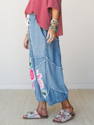 A side-profile shot of relaxed chambray jogger pants with patchwork paneling, frayed seams, and a bold rose appliqué at the hem — styled with a red striped top, stacked turquoise bracelets, and beaded sandals for a mix-it-all boho energy.
