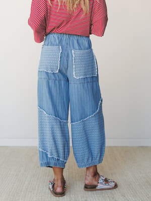 A back-view shot of wide-leg chambray pants with mixed patchwork textures, frayed seam detailing, and an elastic waistband and cuffs — styled with a red striped top and embellished sandals against a clean studio backdrop.