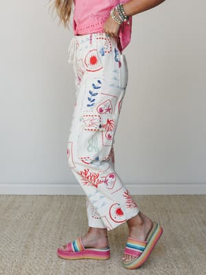 A side-profile shot of cream cargo pants covered in hand-drawn-style ocean and nature motifs — seahorses, coral, waves, botanicals — paired with a pink eyelet crop top, rainbow platform sandals, and a stack of beaded bracelets that somehow pulls the