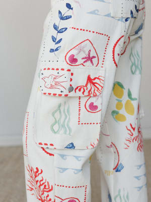 A close-up detail shot of white wide-leg cargo pants covered in a whimsical illustrated print featuring sea creatures, botanicals, hearts, birds, and lemons — the cargo pocket flap snapped shut and outlined in red dotted stitching, the whole thing fe