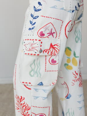 A close-up detail shot of white wide-leg cargo pants covered in a whimsical illustrated print featuring sea creatures, botanicals, hearts, birds, and lemons — the cargo pocket flap snapped shut and outlined in red dotted stitching, the whole thing fe