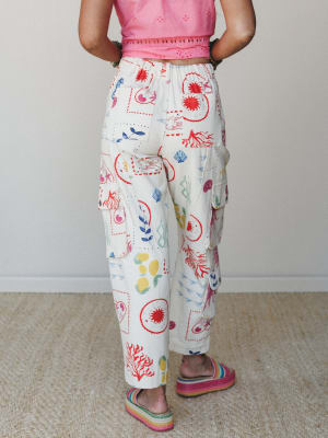 A back-facing shot of wide-leg cargo pants in cream with a hand-drawn nautical and nature print — fish, coral, lemons, botanicals — paired with a pink eyelet crop top and rainbow platform sandals for a look that feels like summer in art class.