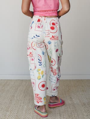 A back-facing shot of wide-leg cargo pants in cream with a hand-drawn nautical and nature print — fish, coral, lemons, botanicals — paired with a pink eyelet crop top and rainbow platform sandals for a look that feels like summer in art class.