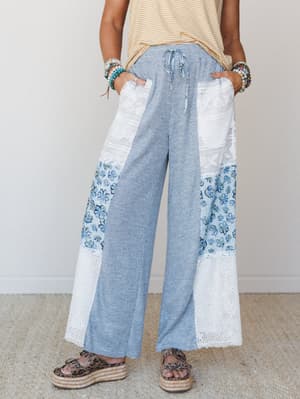 Patchwork wide-leg pants in heathered blue with lace panels and floral print inserts, styled casually with a striped tank, stacked beaded bracelets, and espadrille platform sandals — the kind of outfit that looks effortless but definitely took a seco