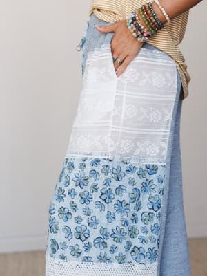 A close-up detail shot of wide-leg pants in a mix of chambray blue, white crochet lace, and a watercolor floral print panel, styled with a mustard stripe tee and a stack of beaded bracelets — the kind of outfit that looks like it was put together eff