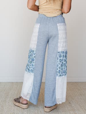 A back-facing model shows off wide-leg patchwork pants in heathered blue, combining floral print panels, lace trim, and crochet inserts — styled with a mustard stripe tee and leopard print espadrille flatforms for an effortlessly collected boho look.