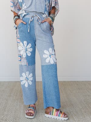 Wide-leg patchwork pants in soft denim blue with lace daisy appliqués paired with a matching top, stacked bead bracelets, and colorful platform sandals — the kind of outfit that's equal parts free-spirited and effortlessly put-together.