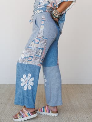 A side-profile shot of wide-leg pants in heathered blue with patchwork fabric panels and daisy appliqués, styled with a floral tie-front top, stacked beaded bracelets, and rainbow woven sandals — the whole look has that effortless, 'I raided a vintag