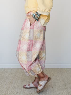 A side-profile shot of relaxed harem-style pants in a pastel paisley patchwork print, styled with a soft yellow sweatshirt, layered bead bracelets, and white buckle sandals — the kind of outfit that feels like a slow Saturday with nowhere to be.