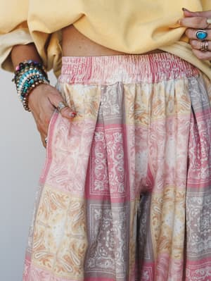 A close-up waist shot of flowy paisley-print pants in soft pink, cream, and golden tones, styled with a knotted butter yellow sweatshirt and layered boho jewelry — the kind of outfit that looks effortless but was definitely thought about.