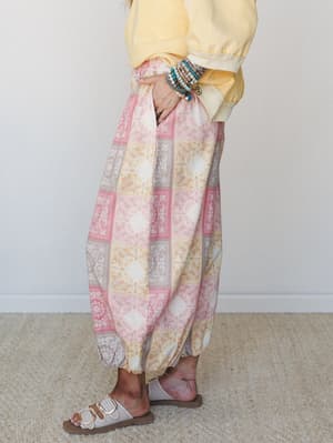 A side-profile shot of harem-style pants in a soft patchwork paisley print — pink, golden yellow, and taupe — styled with a butter yellow sweatshirt, layered bead bracelets, and white buckle sandals for an effortlessly relaxed boho look.