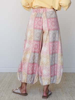 A back-view shot of wide-leg harem-style pants in a soft patchwork bandana print — rosy pinks, warm tans, and dusty lavender — paired with a butter yellow cropped sweatshirt and flat sandals for an effortlessly relaxed boho look.