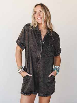 A washed-out black short romper with a relaxed shirt-style silhouette, styled with stacked beaded bracelets and a dainty necklace — the kind of effortless one-and-done outfit that just works.