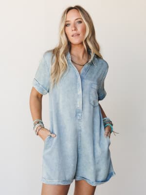 A light wash denim-look romper styled with stacked beaded bracelets and a dark beaded necklace — effortless and a little lived-in, like she just threw it on and somehow nailed it.