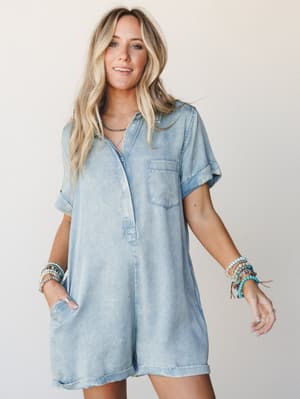 A light wash chambray-style romper worn with stacked beaded bracelets and a relaxed smile — the kind of outfit that works for a farmers market run or a spontaneous road trip stop.