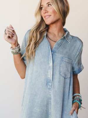 A relaxed chambray-style shirt dress or romper in a soft acid-washed blue-gray, styled with stacks of beaded bracelets and a delicate dark bead necklace — the kind of effortless outfit that looks like it asked zero questions this morning.