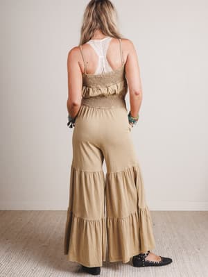 Woman wearing a beige tiered jumpsuit with a white lace top underneath, standing against a plain background.
