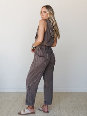 A model shot from behind looking over her shoulder, wearing a washed brown sleeveless hooded jumpsuit with exposed seam detailing and a relaxed straight-leg silhouette — effortlessly lived-in and ready for wherever the day goes.