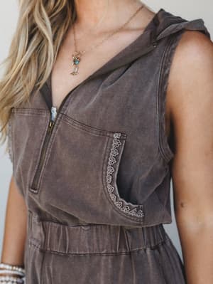 A close-up torso shot of a washed brown sleeveless hooded jumpsuit with a half-zip front, lace-trimmed chest pocket, and elastic waist — styled with layered gold necklaces and pearl bracelets for that effortless, lived-in boho feel.
