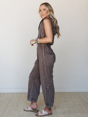 A washed brown sleeveless jumpsuit with lace trim detail and a relaxed jogger silhouette, styled with stacked boho bracelets and white sandals — the kind of one-and-done outfit that still looks like you tried.