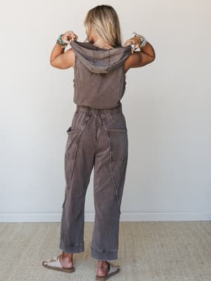 Shot from the back, a model pulls up the hood of a washed brown sleeveless hooded jumpsuit with exposed seam detailing and wide-leg pants — styled with beaded bracelets and sandals for an effortlessly lived-in feel.