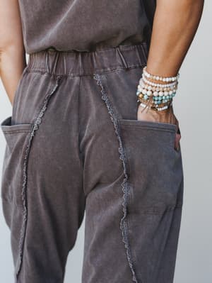 A close-up detail shot of a washed brown jumpsuit, shot from the back — the lace trim seaming and patch pockets do the quiet talking while a stacked bracelet situation on the wrist adds just the right amount of personality.