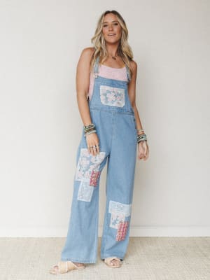 Wide-leg denim overalls with mixed lace and fabric patchwork details, styled over a pink lace tank with stacked bracelets and sandals — the kind of outfit that looks like it was put together effortlessly but definitely wasn't.
