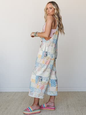 A wide-leg patchwork denim overall jumper in soft pastel florals and quilted blocks, styled over a pink lace bralette with rainbow platform sandals and stacked bead bracelets — the kind of outfit that looks like a vintage quilt came to life in the be