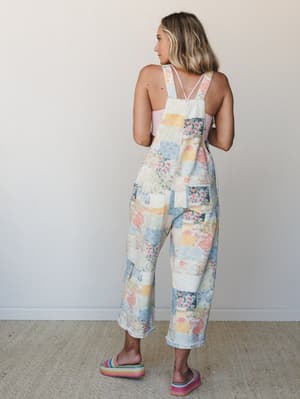 A model shot from behind showing wide-leg patchwork denim overalls in a soft pastel quilt-like print, styled with a pink cami underneath and rainbow platform slides — easy, expressive, and the kind of outfit that makes strangers ask where you got it.