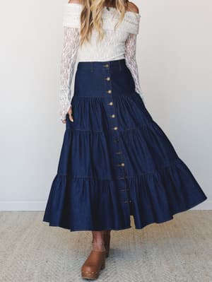 A tiered dark denim maxi skirt with brass button-front closure, styled with an off-shoulder lace top and tan clogs — the kind of outfit that makes a grocery run feel like a whole moment.