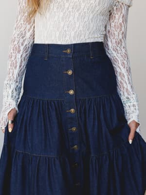 A tiered dark denim maxi skirt with brass button-front closure paired with a sheer white lace long-sleeve top — the kind of outfit that somehow works for both a farmers market and a date night.
