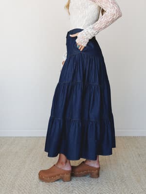 A dark denim tiered maxi skirt styled with a sheer lace long-sleeve top and brown studded clogs — the kind of outfit that feels equally at home at a farmers market or a casual dinner out.