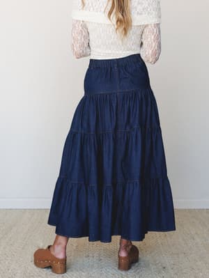 Shot from behind, a tiered dark denim maxi skirt pairs with a cream lace long-sleeve top and brown wood-heel clogs — the kind of outfit that feels effortless but clearly thought through.