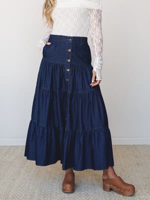 A dark denim tiered maxi skirt with button-front detail is styled with a sheer lace long-sleeve top and brown studded clogs — the kind of outfit that feels both rooted and a little romantic.