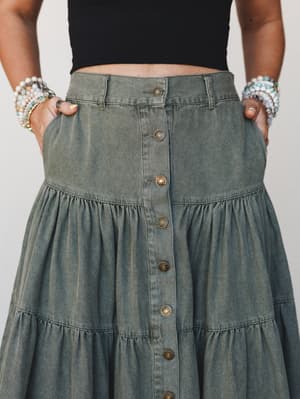 A close-up mid-shot of an olive-washed denim tiered maxi skirt styled with a black crop top and stacked beaded bracelets — the kind of skirt that makes a farmer's market run feel like a whole moment.