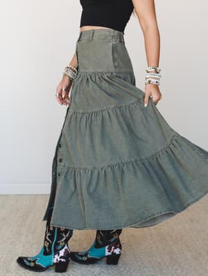 A side-profile shot of a tiered denim maxi skirt in washed olive, styled with a black crop top and bold embroidered turquoise cowboy boots — the kind of outfit that makes a Saturday feel intentional.