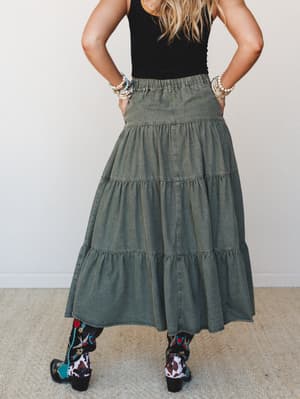 A back-view shot of a tiered olive maxi skirt styled with a black tank and bold embroidered cowboy boots — the kind of outfit that's equally at home at a weekend market or a dusty outdoor concert.