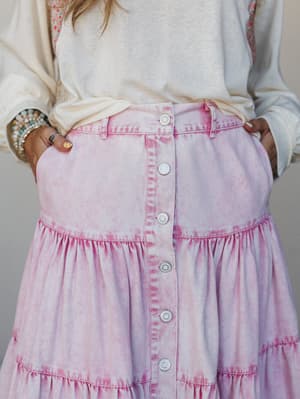 A close-up of a pink tie-dye denim tiered maxi skirt styled with a cream sweatshirt and layered bracelets — the kind of piece that makes a casual Saturday feel intentional.