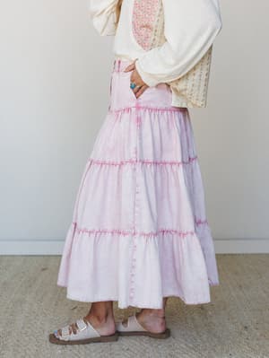 A side-profile shot of a tiered acid-wash maxi skirt in soft pink, styled with a cream oversized top and floral panel detail, sandals, and a turquoise ring — casual and a little dreamy.