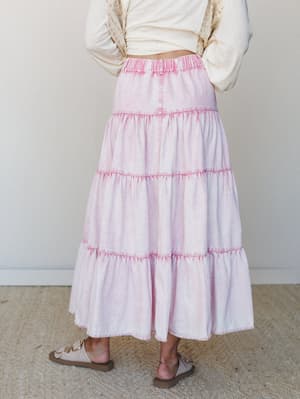 A back-view shot of a tiered maxi skirt in acid-washed pink denim, paired with a knotted cream top and neutral sandals — soft, lived-in, and effortlessly boho.